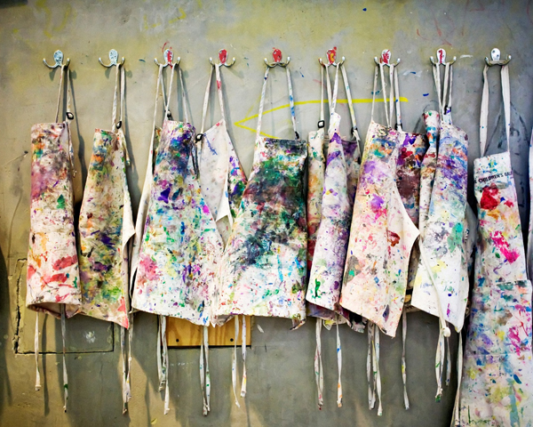 Eight hooks with mutil-coloured paint-stained aprons hanging from them.