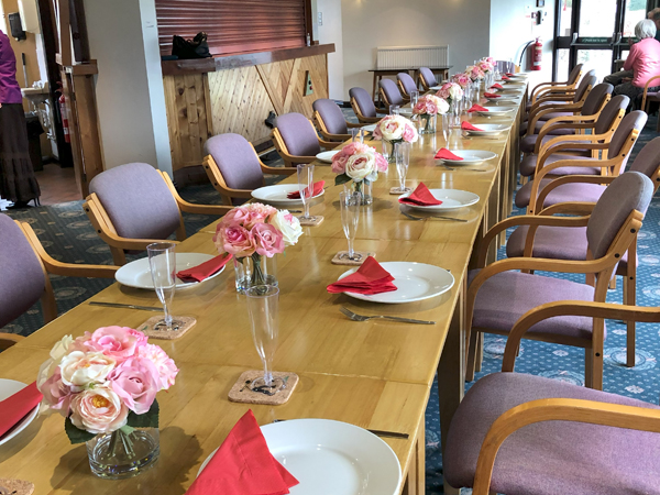 A long table set with pick and white flower centrepieces, plates, red napkins, cutlery and flutes.