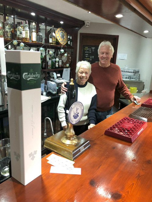 Woman and man behind the bar, looking at the camera and smiling.