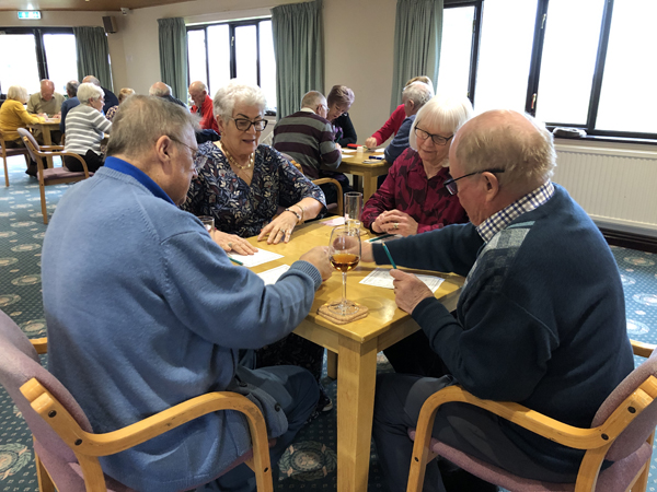 Picture taken from the back of the Clubhouse. Four tables of four people playing Beetle. In the foreground, Kevin, Pat, Karen and Ian.