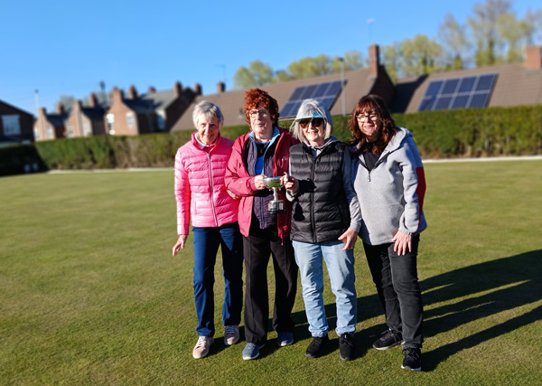 Four women standing on a green, posing with the Pairs Trophy.