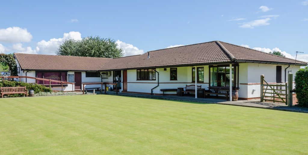 The Clubhouse. Picture taken from the green. A white building in an L shape. The windows and doors are brown. The roof tiles are reddish-brown. There are benches under the eaves on the right hand side. A slope (with railings) down to the front door on the left-hands side.