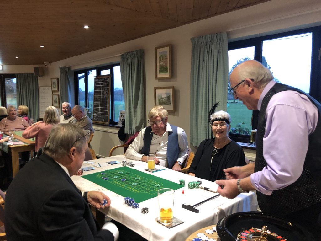 Two men and a women seated around a roulette table. One man standing taking bets.
