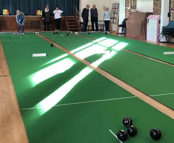 Looking across the three mats at Claremount Bowling Club. Bowlers are at the far end. Rays of sunshine bisect the mats.