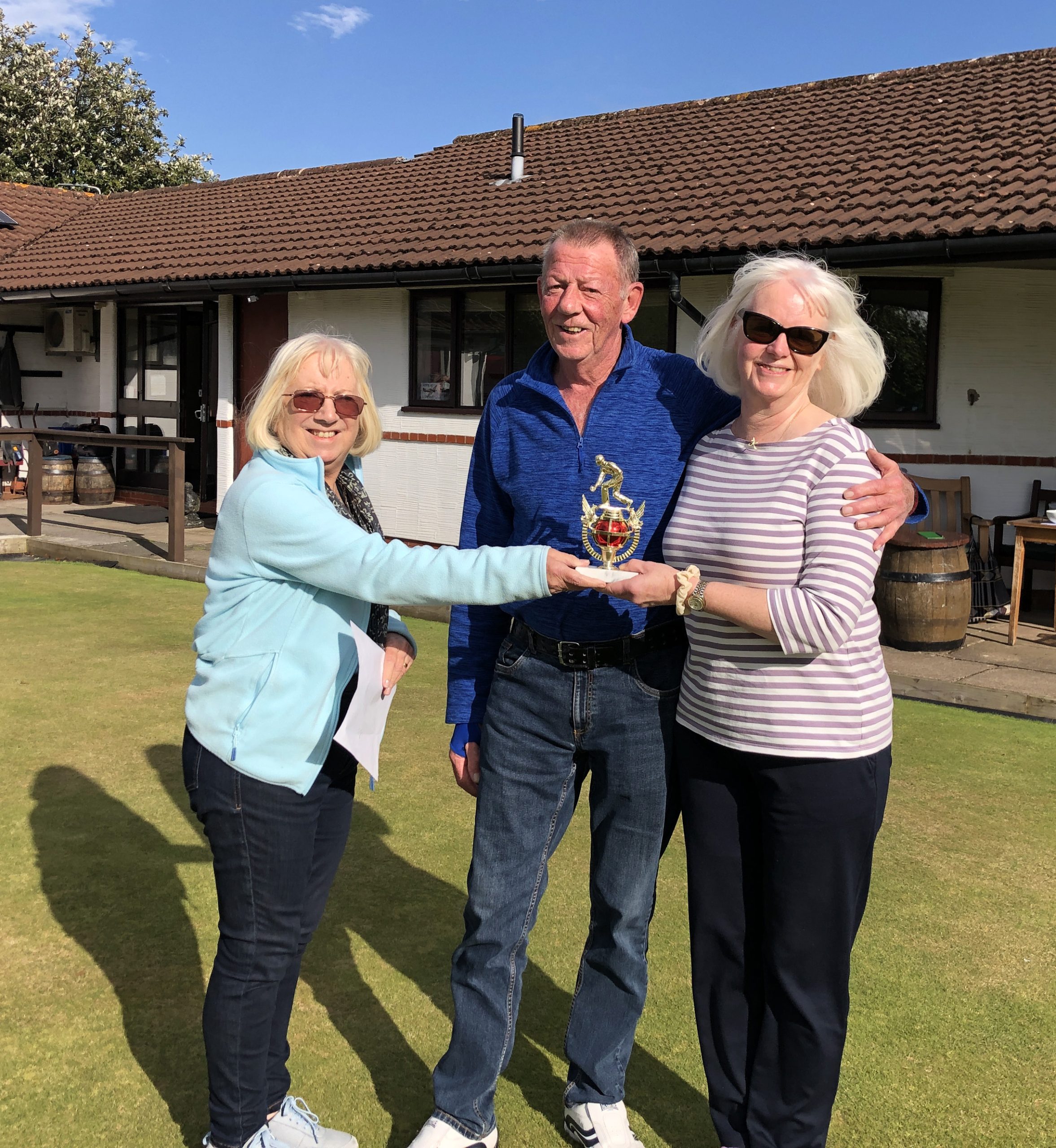 Two women and a man standing on the green. The man is in the middle and has his arm around the shoulder of the woman on the right. The woman on the left is handing over the small trophy.