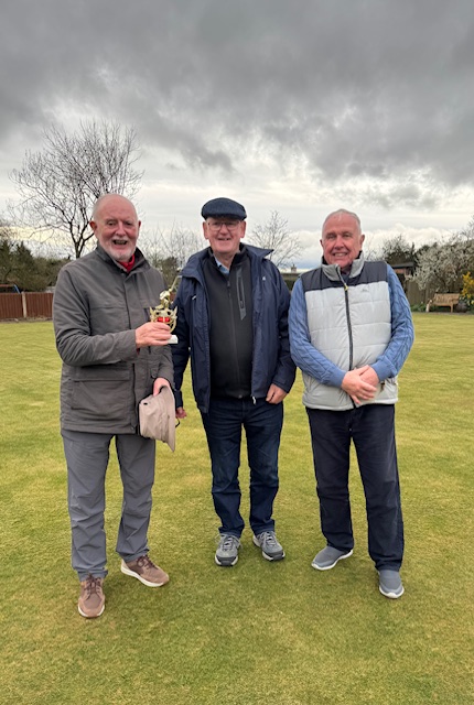 Three men standing on the green, posing with the small trophy.