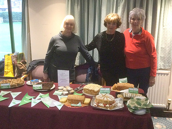 Karen, Jackie & Margaret standing behind a table with cakes for sale.