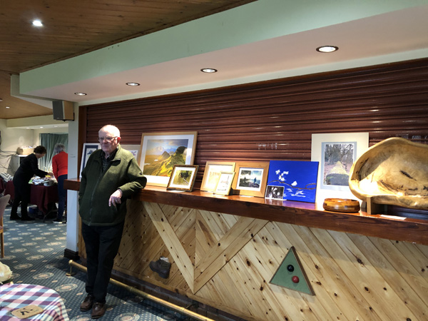 Barry standing by the bar next to his photograph of a kingfisher. Paintings are also displayed on the bar.