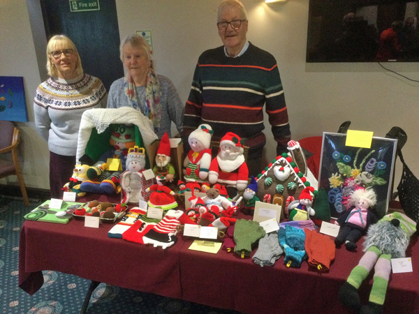 Chris, Maggie & Pat standing behind a table, displaying Maggie's knitting projects, Christmas themed.