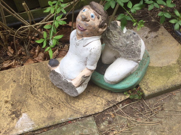 A ceramic figure of a male bowler, dressed in white, holding a black bowl. The figure is broken into two pieces, just below its waist. It has been placed on a flagstone in the garden.