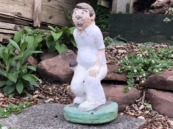 A ceramic figure of a male bowler, dressed in white, holding a black bowl. The figure has been placed on a flagstone in the garden.
