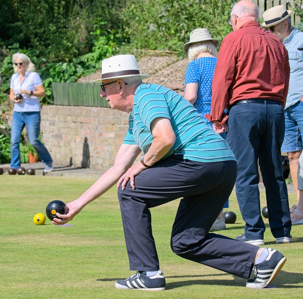 A man bending down and delivering a bowl. Men and women in the background looking at an end to determine who has won. Woman in the background in another game, watching.