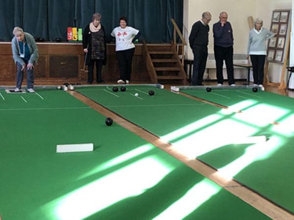 Looking across the three mats at Claremount Bowling Club. Bowlers are at the far end. Rays of sunshine bisect the mats.