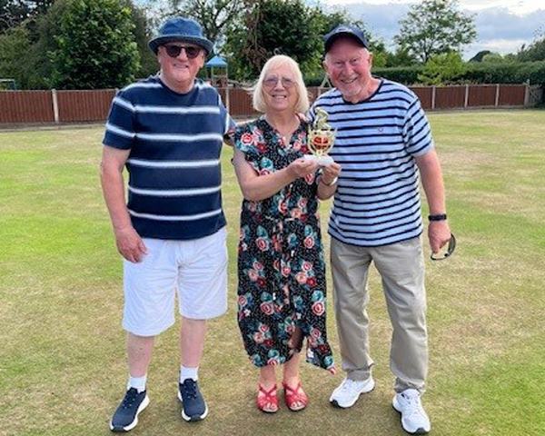 On the green, two men standing either side of a woman who is holding a small trophy.