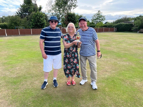 On the green, two men standing either side of a woman who is holding a small trophy.