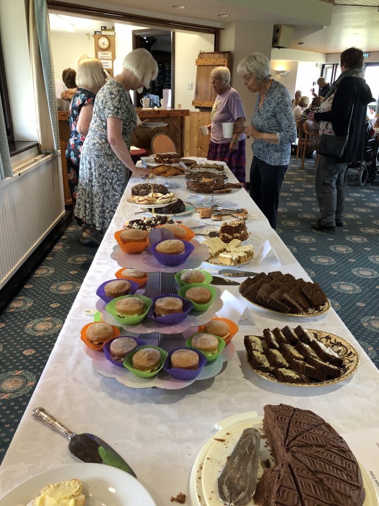 Cakes set out on long tables. A Club member serving and people in the background waiting for drinks.
