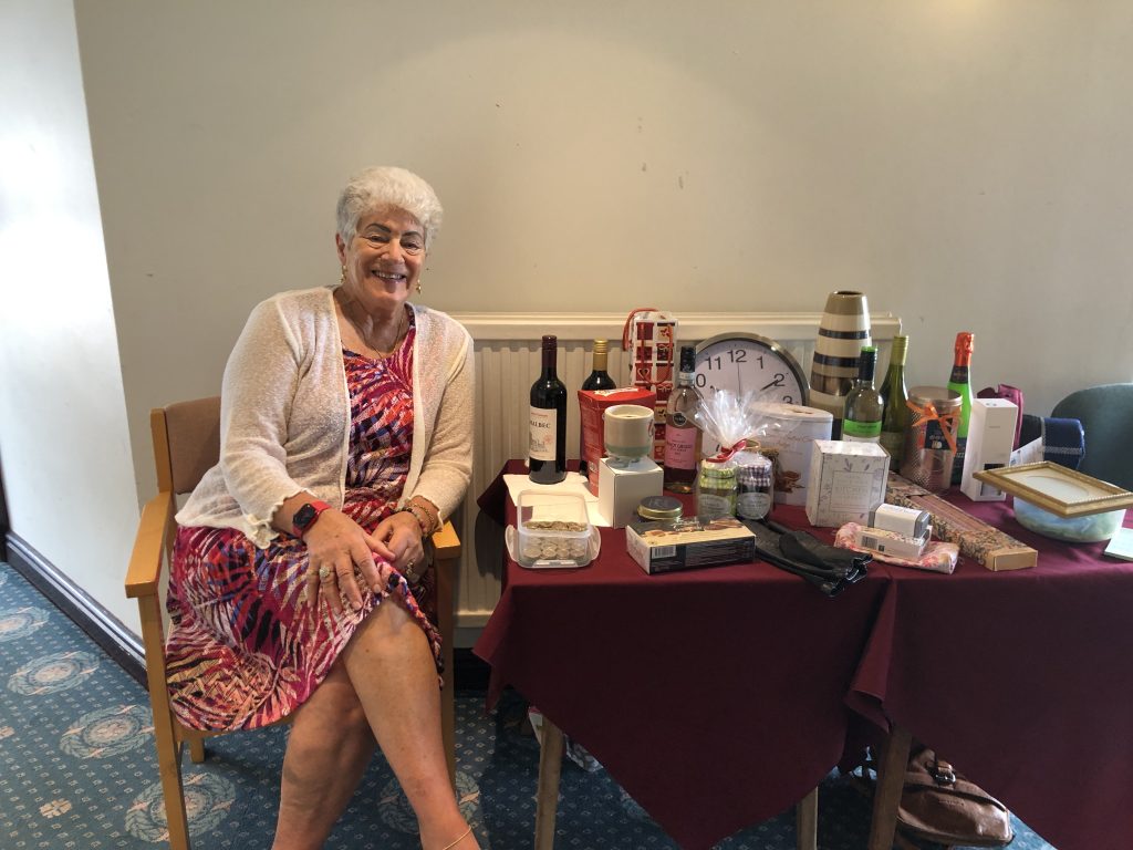 Pat sitting at the raffle table with the many prizes - bottles of wine, candles, a clock, vases, and edibles.