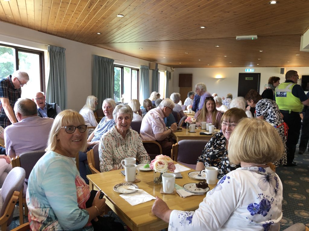 The Clubhouse full of people sitting at tables with tea/coffee and cake.