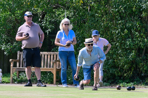 Two women and two men on the green. One man is the process of delivering a wood, the others are standing behind him, watching.