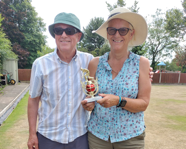A man and a woman standing on the green, smiling at the camera. The woman is holding the small trophy.