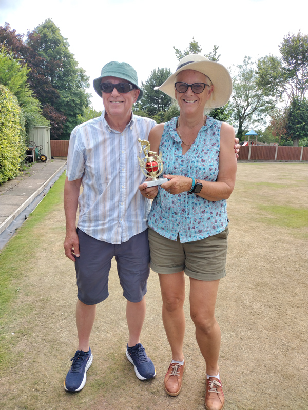 A man and a woman standing on the green, smiling at the camera. The woman is holding the small trophy.