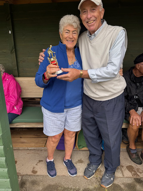 A woman and a man standing in the cover of the Bowls shelter. They are posing with the small trophy.