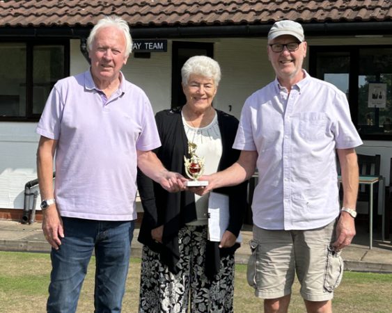 Two man standing either side of a woman on the green. They are holding the small trophy.