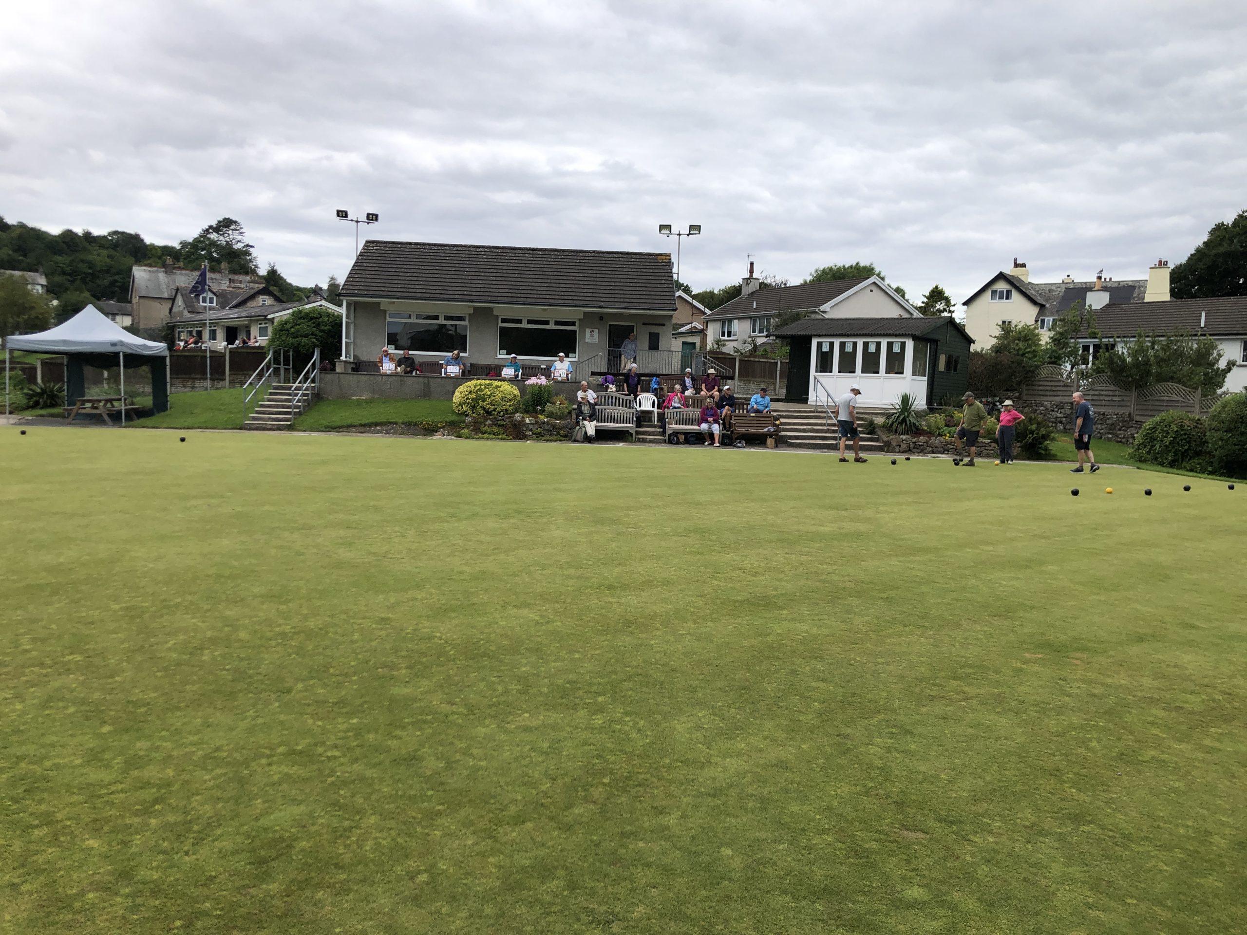 Arnside Bowling Club The Clubhouse and small conservatory are at the back. Bowlers are on the green and others are sitting watching. Markers are sitting up by the Clubhouse.
