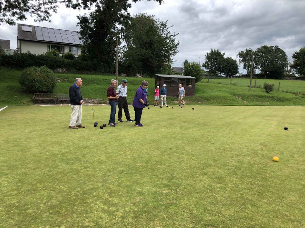 Men and women on the green In the foreground a woman is getting ready to bowl watched by three men. In the background, two men and woman are assessing the points in another game.