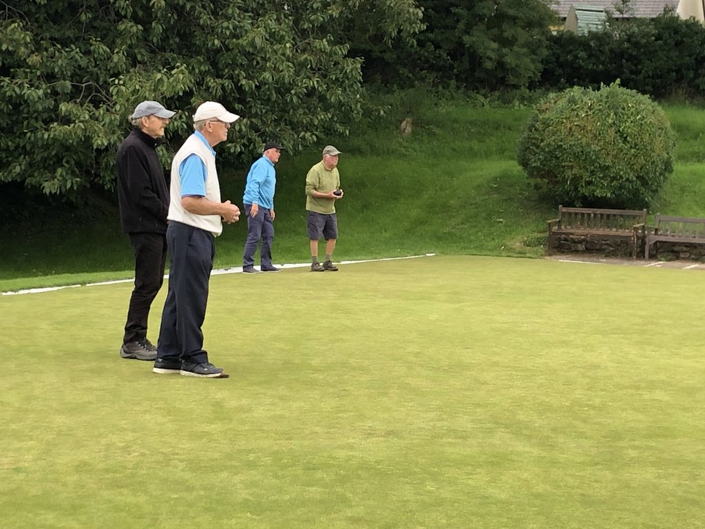 Four men on the green at Arnside Bowling Club. Two pairs - the front man in each case is holding a bowl and getting ready to bowl.