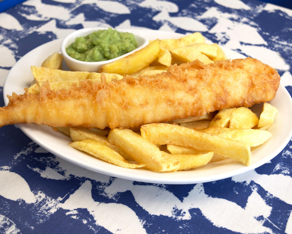 Fish and chips and mushy peas on a white plate. The plate set on a blue tablecloth with a white fish motif.