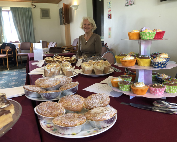 Lindy sitting behind a table with cakes on plates and on a three tier cake stand.