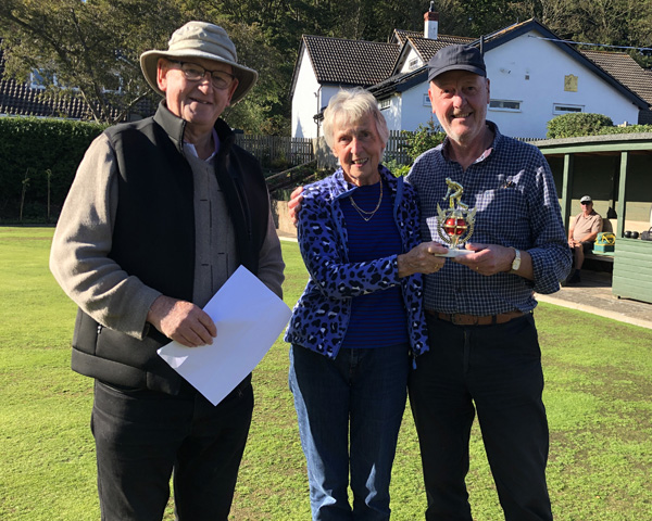 A woman standing between two men on the green. The woman and the man to right are holding the small trophy.