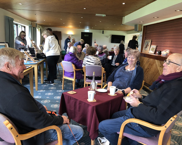 People sitting at tables in the Clubhouse, drinking tea and coffee and eating cake. Craft tables down the left-hand side.