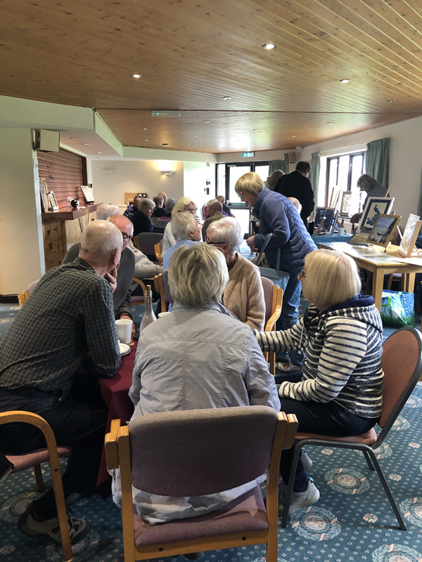 People sitting at tables in the Clubhouse, chatting, drinking tea and coffee and eating cake. Tables with crafts down the right hand side.