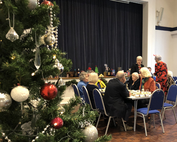 A view of the Hall. In the right hand foreground part of a Christmas tree. People sitting in tables and chairs.