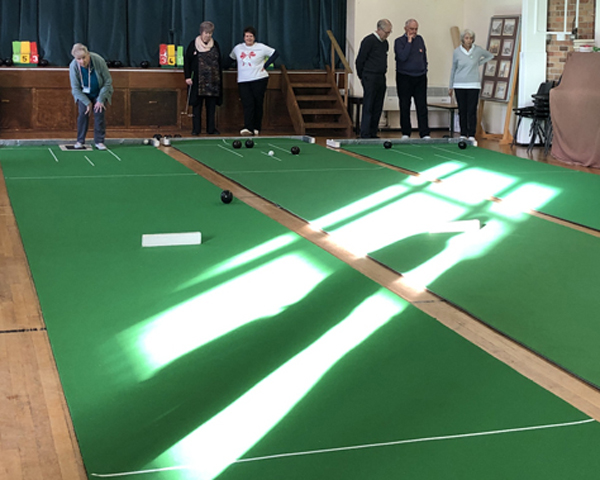 Looking across the three mats at Claremount Bowling Club. Bowlers are at the far end. Rays of sunshine bisect the mats.
