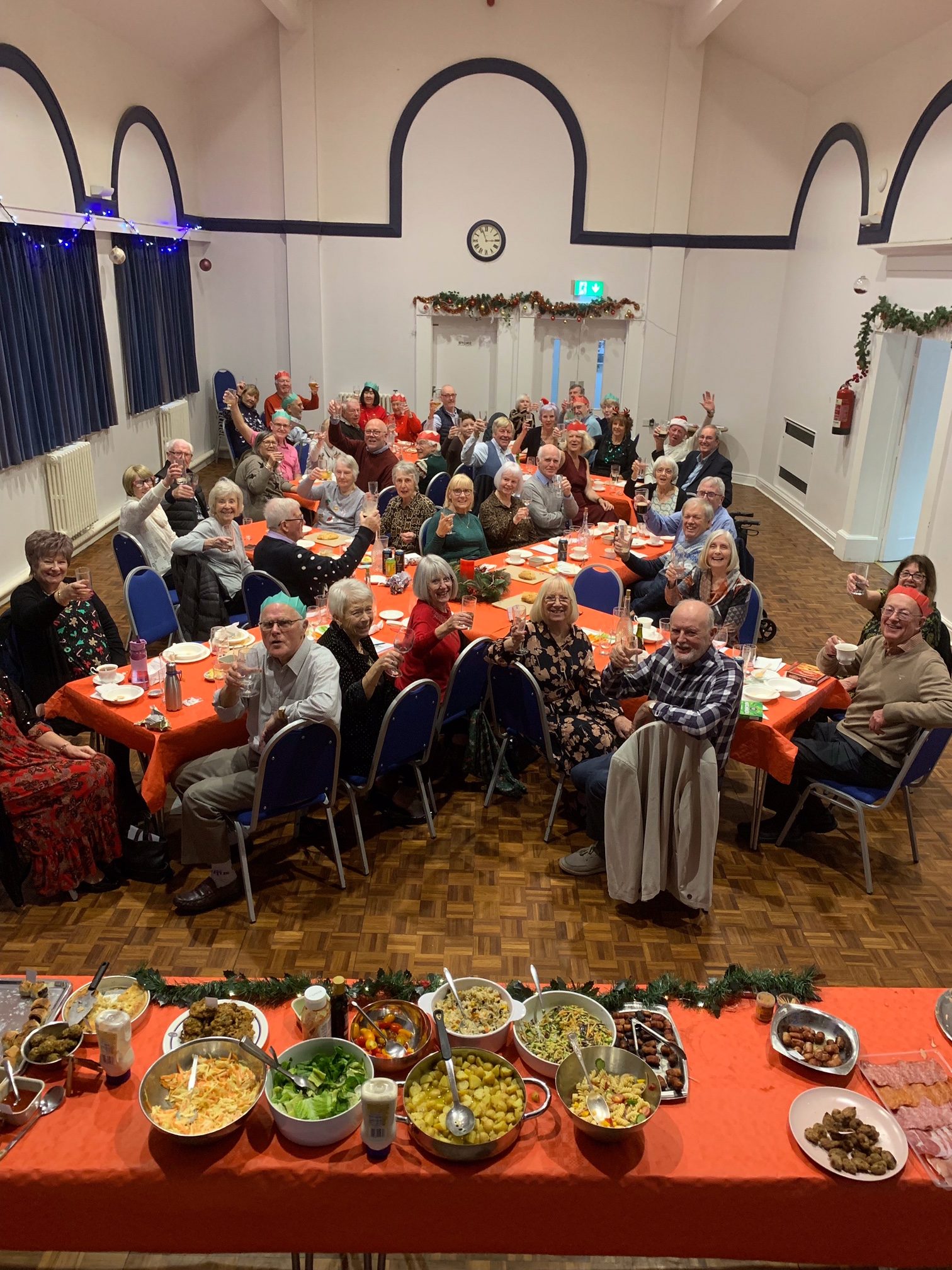 Picture taken from the stage. People sitting at the tables, looking up at the stage, smiling and raising their glasses. Food table in the foreground.