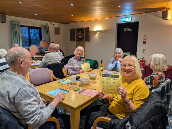 People sitting at a table, smiling at the camera. Two women holding up bingo cards.