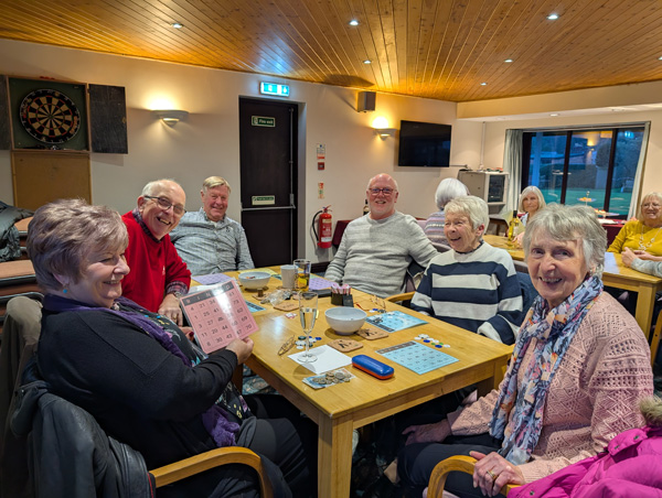 People sitting at a table, smiling at the camera. One woman holding up a bingo card.