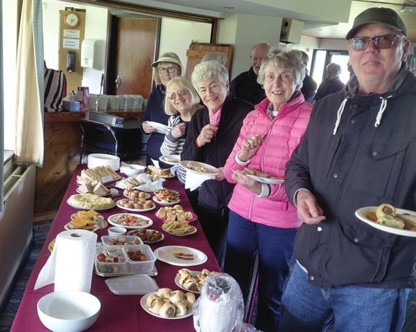 People standing by a long table in the Clubhouse with food on paper plates. All smiling at the camera.