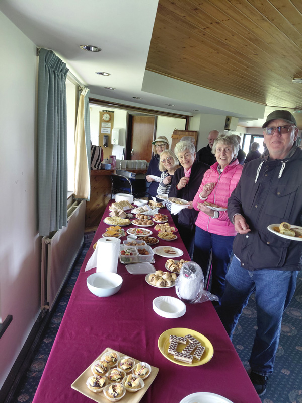 People standing by a long table in the Clubhouse with food on paper plates. All smiling at the camera.