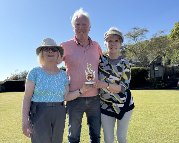 A man standing between two women on the green. The women are holding the small trophy.