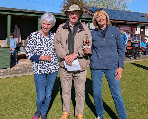 A man standing between two women on the bowling green, smiling at the camera. The woman on the right is holding the small trophy.