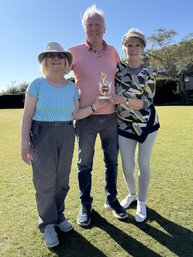 A man standing between two women on the green. The women are holding the small trophy.