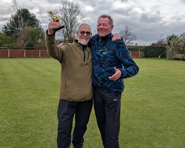 Two men standing on the green, smiling at the camera, with arms around each other's shoulders. The man on the left is holding up the small trophy.