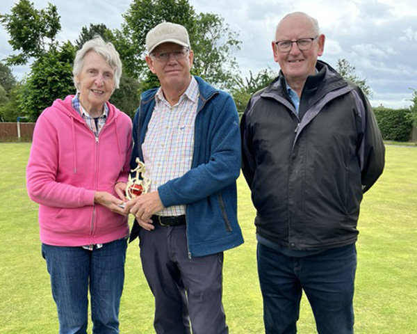 A woman and two men standing on the green. The woman and man on the left are holding the small trophy.