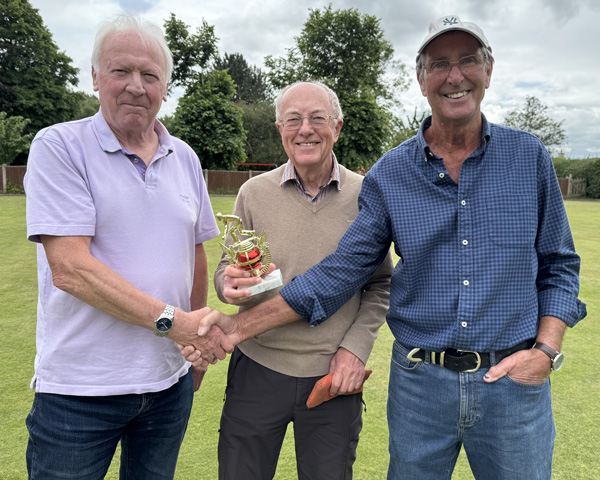 Three men standing on the green. The two on the outside are shaking hands. The one in the middle holds the small trophy.