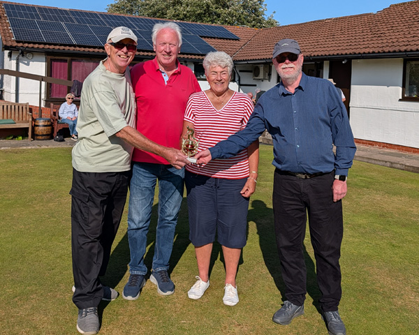 Three men and one woman standing on the green. The men on the right and left are holding the small trophy.