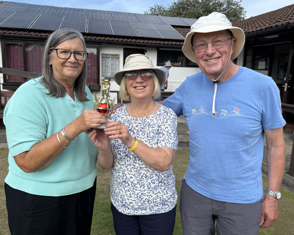 Two women and one man standing on the green. The two women are holding the small trophy.